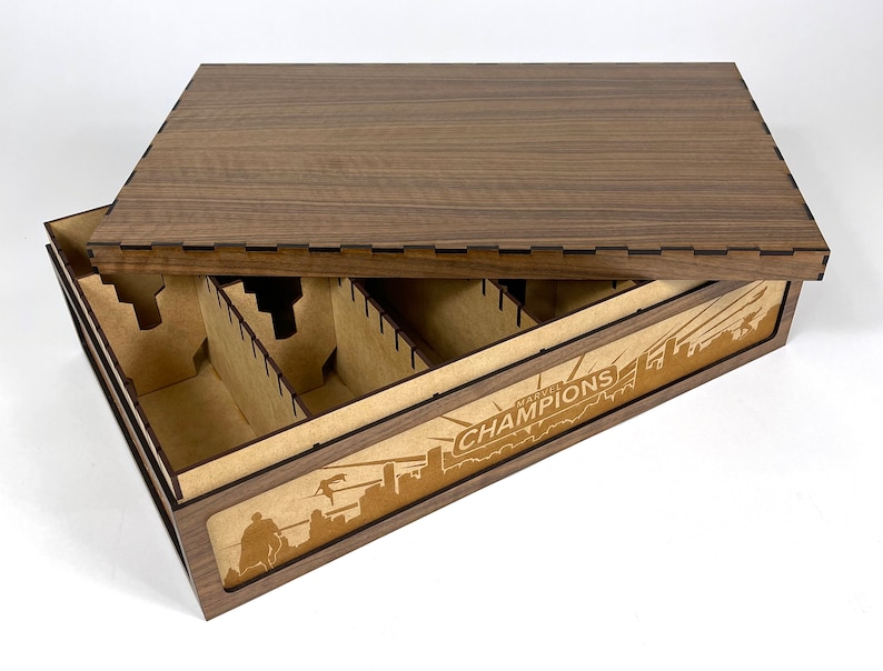 May include: A wooden box with a lid, designed to store and organize a board game. The box has a brown wood finish and features a laser-engraved design of the Marvel Champions logo and a cityscape. The interior of the box is divided into compartments for storing game components.