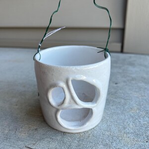 May include: A white ceramic planter with three oval cutouts and a green wire handle. The planter has a textured surface and a heart-shaped handle. Ideal for small plants or succulents.