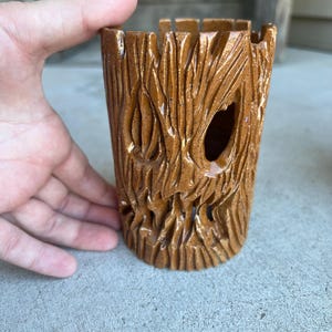 May include: A brown ceramic planter with a tree bark texture. The cylindrical planter has a carved design with a face-like appearance and a large opening. The planter is held by a hand.
