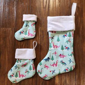 May include: Three Christmas stockings with a dinosaur theme. The largest stocking is teal blue with white trim and features a variety of dinosaurs, trees, and presents. The two smaller stockings are teal blue with white trim and feature a variety of dinosaurs, trees, and presents.