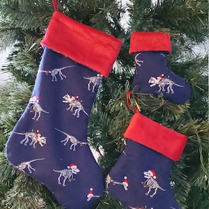 May include: Three Christmas stockings with a blue background and a red cuff. The stockings are decorated with a white dinosaur skeleton pattern. The dinosaur skeletons are wearing Santa hats.