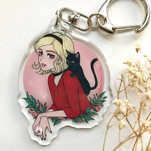 May include: A pink and white round acrylic keychain featuring a cartoon illustration of a woman with blonde hair and a black cat. The woman is wearing a red sweater and a black choker. The text "Moondust Ore" is written on the keychain.