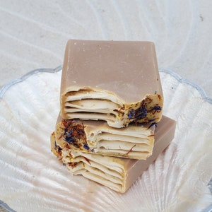May include: Three layers of handmade soap with a light brown color and a beige and brown swirl pattern. The soap is decorated with dried flowers and petals in shades of blue, purple, and orange.