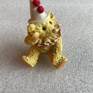 May include: A small, yellow teddy bear figurine dressed as a clown. The bear wears a white hat with two red pom-poms and a ruffled collar with red trim. Its feet and nose are orange.