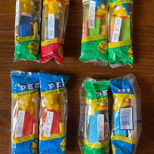 May include: Ten individually packaged Simpsons character PEZ dispensers. Each dispenser is yellow with a blue, green, or red body. The packaging is clear plastic with a blue, green, or red background. The packaging includes the PEZ logo and the text "Candy & Dispensers".
