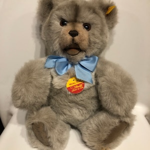 May include: A gray plush teddy bear with a blue bow tie. The bear has a button in its ear and a tag that reads "Steiff Knopf im Ohr".
