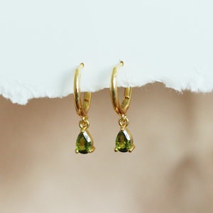 May include: A pair of gold hoop earrings featuring small, dangling, pear-shaped green gemstones. The earrings are suspended from a white, textured edge, showcasing the polished gold metal hoops and the faceted, vibrant green teardrop stones.