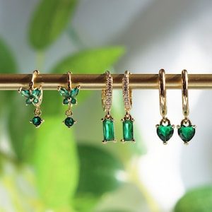 May include: A set of four gold hoop earrings with dangling charms. The charms are a butterfly, a rectangle, and two hearts. All of the charms are adorned with green gemstones.