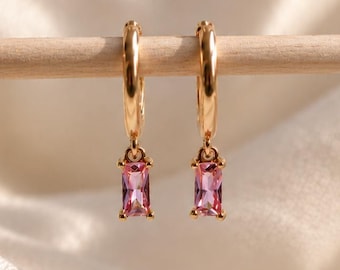 May include: A pair of gold hoop earrings, each featuring a single dangling pink baguette-cut gemstone. The earrings are suspended from a thin wooden rod, showcasing the metallic gold finish and the clear, vibrant pink color of the rectangular stones.