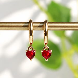 May include: A pair of gold hoop earrings, each featuring a small, faceted, heart-shaped red gemstone charm dangling from the bottom. The earrings are suspended from a thin, horizontal gold-toned metal bar.