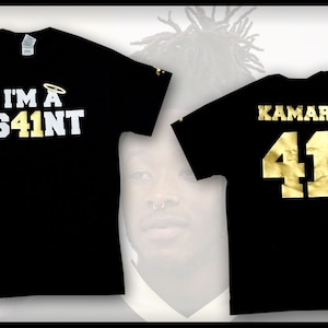 Peut inclure: T-shirt noir avec inscription dorée. Le devant du t-shirt dit "I'm a S41NT" avec un halo au-dessus du 41. Le dos du t-shirt dit "KAMARA 41".