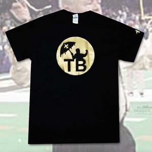 May include: Black t-shirt with a gold circle graphic featuring a silhouette of a person holding an umbrella with a fleur-de-lis on top. The circle also contains the letters "TB".