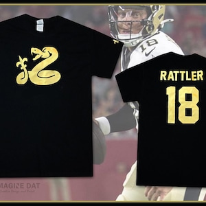 Puede incluir: Dos camisetas negras con letras y gráficos dorados. La primera camiseta presenta un gráfico de serpiente dorada con una flor de lis. La segunda camiseta tiene el texto "RATTLER" y el número "18" en dorado.
