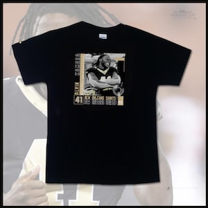 Peut inclure: T-shirt noir avec une image en niveaux de gris d'Alvin Kamara, joueur de football américain, les bras croisés. Le t-shirt affiche "ALVIN KAMARA" et "41 NEW ORLEANS SAINTS" en or et blanc. La marque "IMAGINE DAT" est imprimée en haut à droite.