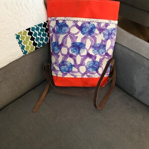 Puede incluir: Un bolso tote de lona con la parte superior e inferior de color naranja brillante, y un panel central blanco con un patrón abstracto azul y morado. El bolso tiene correas de cuero marrón.