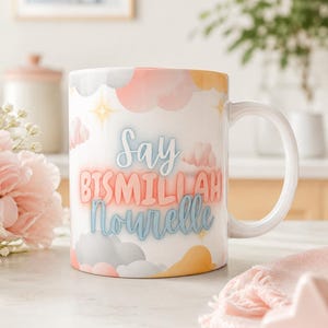Peut inclure: Un mug en céramique blanche orné d'un motif de nuages aux tons pastel rose, jaune et gris, agrémenté de petites étoiles jaunes. Le texte "Say BISMILLAH Nourelle" est imprimé sur le devant dans une combinaison de lettres cursives et capitales.