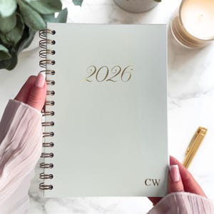 Può includere: Un'agenda verde chiaro a spirale con l'anno "2026" in oro. Le iniziali "CW" sono in basso a destra. L'agenda è tenuta da una persona con unghie curate.