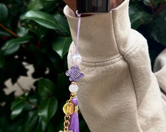 May include: A black lipstick tube with a decorative purple tassel. The tassel features a gold butterfly charm, a purple knot charm, a white bead, and a pearl bead. The tassel is attached to the lipstick tube with a thin, light purple string.