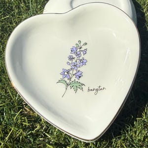 May include: Heart-shaped, off-white ceramic dish with a gold rim. The dish features a delicate illustration of purple flowers and green leaves, with the word "bangtan" in black script. Another heart-shaped dish is visible in the background.