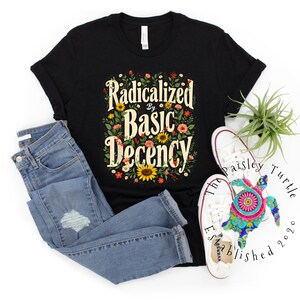 Puede incluir: Camiseta negra con la frase "Radicalized by Basic Decency" en un diseño floral. La camiseta se combina con vaqueros azules y zapatillas blancas. La imagen también incluye una tortuga decorativa y una pequeña planta.