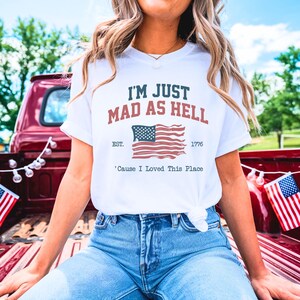 Puede incluir: Camiseta blanca con el texto "I'M JUST MAD AS HELL" en azul marino y rojo, junto con un gráfico de la bandera estadounidense. La camiseta también incluye el texto "EST. 1776" y "'Cause I Loved This Place". La persona lleva vaqueros.