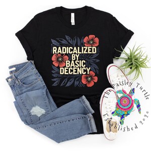 Puede incluir: Camiseta negra con el texto "RADICALIZED BY BASIC DECENCY" en un diseño floral. La imagen incluye vaqueros azules y zapatillas blancas. También hay una pequeña planta y una tortuga decorativa.
