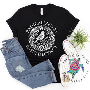 Puede incluir: Camiseta negra con el texto "RADICALIZED BY BASIC DECENCY" en un diseño floral blanco con un pájaro. La imagen incluye vaqueros azules y zapatillas blancas. Una pequeña planta y una tortuga decorativa también están en la imagen.