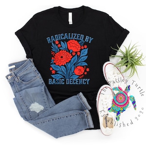 Puede incluir: Camiseta negra con el texto "RADICALIZED BY BASIC DECENCY" en azul. La camiseta presenta un diseño floral con flores rojas y hojas azules. También hay vaqueros azul claro y zapatillas blancas en la imagen.