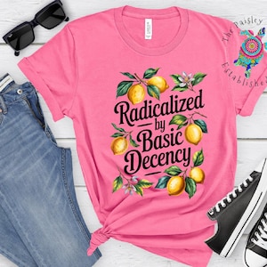 Puede incluir: Una camiseta rosa con el texto "Radicalized by Basic Decency" e ilustraciones de limones. La camiseta se combina con vaqueros azules, gafas de sol negras y zapatillas negras.