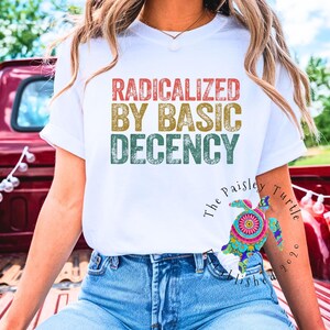 Puede incluir: Camiseta blanca con el texto "RADICALIZED BY BASIC DECENCY" en rojo, dorado y verde. La camiseta se usa con vaqueros azules. La imagen incluye banderas americanas y una caja de camión roja en el fondo.