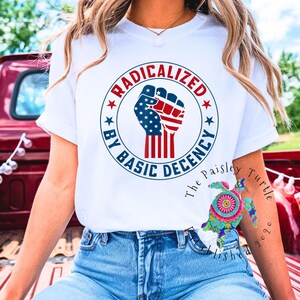Puede incluir: Camiseta blanca con el texto "RADICALIZED BY BASIC DECENCY" que rodea un diseño de puño en rojo, blanco y azul. La camiseta se usa con vaqueros azules. Banderas americanas visibles en el fondo.