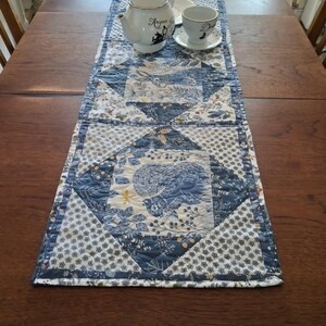 May include: A rectangular quilted table runner featuring a blue and white fabric pattern with illustrations of squirrels and rabbits. The runner is decorated with geometric patchwork squares and floral motifs, displayed on a wooden table surface.