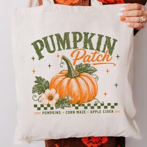 Peut inclure: Un sac fourre-tout en toile couleur crème avec une impression graphique portant le texte PUMPKIN PATCH. Le motif comprend une grosse citrouille orange, des feuilles vertes, une petite fleur blanche et une bordure à carreaux verts avec les mots PUMPKINS, CORN MAZE, APPLE CIDER.