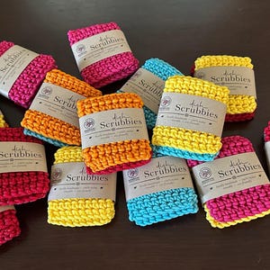 Puede incluir: Una colección de estropajos de cocina hechos a mano y coloridos en varios tonos de rosa, amarillo, naranja, rojo y azul. Cada estropajo está envuelto con una etiqueta de papel que dice "dish Scrubbies". Los estropajos están dispuestos sobre una superficie marrón oscuro.