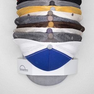 May include: A vertical stack of twelve baseball caps in various colors including white, blue, red, black, gray, yellow, and brown, held by a white wall-mounted display bracket featuring the text DOMEDOCK with a semi-circular icon above it.