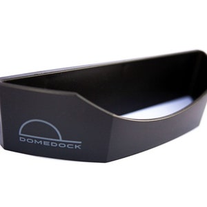 May include: A black, semi-circular plastic storage accessory featuring a flat back and a curved front opening. The word DOMEDOCK is printed in light gray on the front left side, accompanied by a simple semi-circular graphic icon above the text.