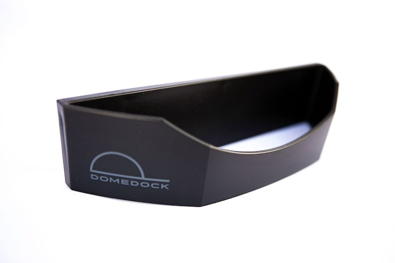 May include: A black, semi-circular plastic storage accessory featuring a flat back and a curved front opening. The word DOMEDOCK is printed in light gray on the front left side, accompanied by a simple semi-circular graphic icon above the text.