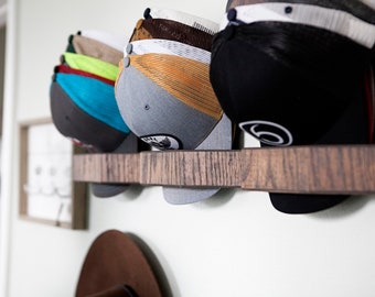 May include: A horizontal wooden wall-mounted rack holding three stacks of baseball caps. The caps feature various colors including black, grey, orange, blue, green, and tan, with mesh back panels and circular patches on the front panels of the hats.