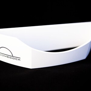 May include: A white, open-top, boat-shaped container with a curved front edge. The side features a black logo consisting of a semi-circle above a horizontal line and the text DOMEDOCK printed in a clean, sans-serif font on the white surface.