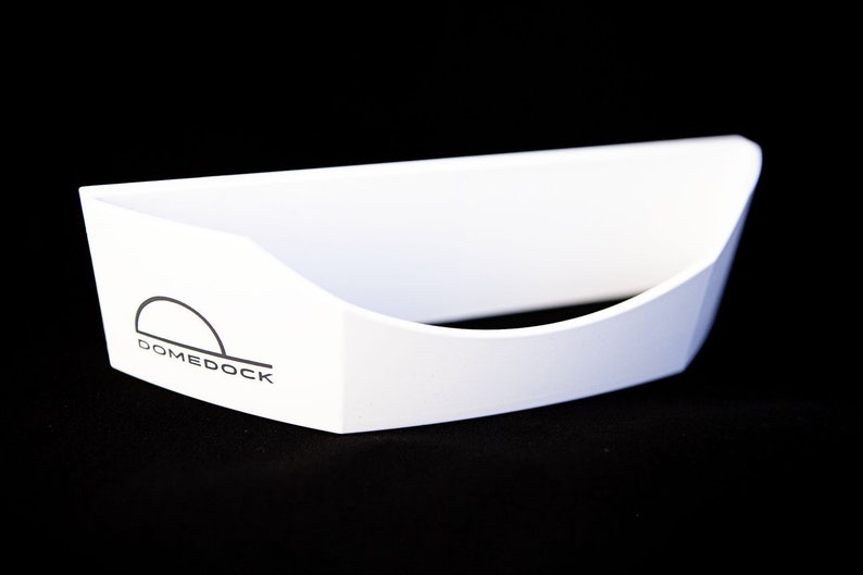 May include: A white, open-top, boat-shaped container with a curved front edge. The side features a black logo consisting of a semi-circle above a horizontal line and the text DOMEDOCK printed in a clean, sans-serif font on the white surface.
