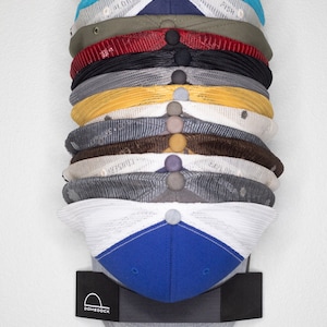 May include: A vertical stack of twelve baseball caps in various colors including white, blue, olive, red, black, gray, yellow, brown, and navy, held by a black DomeDock wall mount featuring a white logo with a hat icon and the text DomeDock.
