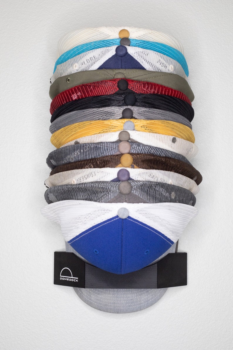 May include: A vertical stack of twelve baseball caps in various colors including white, blue, olive, red, black, gray, yellow, brown, and navy, held by a black DomeDock wall mount featuring a white logo with a hat icon and the text DomeDock.