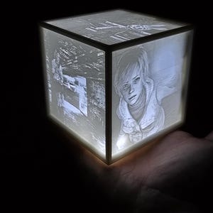 May include: A white cube-shaped night light with a 3D image of a person's face on one side. The light is on and illuminating the image.