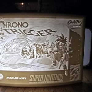 Può includere: Una tazza bianca in ceramica stampata in 3D con un bordo dorato. La tazza presenta un design della copertina del videogioco Chrono Trigger. Il design include il titolo del gioco, i personaggi e il logo di Super Nintendo Entertainment System. La tazza è su una superficie di legno.