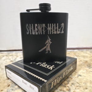 May include: Black hip flask with the text "SILENT HILL 2" and a character graphic. The flask is sitting on top of a black box that says "Hip Flask" and "Size: 6oz".