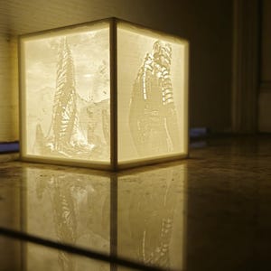 May include: A white cube-shaped lamp with a 3D printed design of a sci-fi character and a tall structure on the sides. The lamp is lit and casting a warm glow.