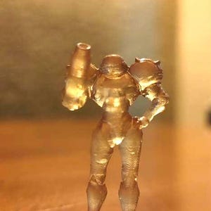 May include: A translucent gold-colored plastic figurine of a female character from a video game. The figurine is standing with one arm raised and holding a weapon.