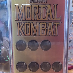Puede incluir: Un llavero rectangular con el texto "MIDWAY MORTAL KOMBAT" en letras doradas sobre un fondo dorado y rojo. El llavero tiene seis botones circulares negros. El llavero está encerrado en plástico transparente.