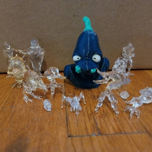Op de afbeelding: Een verzameling van doorzichtige plastic figuurtjes, waaronder een blauw en groen alien-achtig figuurtje, gerangschikt op een houten oppervlak.