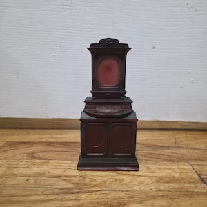 Puede incluir: Un pequeño mueble de madera marrón oscuro con una parte superior decorativa y un interior rojo. El mueble tiene dos puertas en la parte inferior y se asienta sobre una base rectangular. El fondo es una pared blanca y un suelo de madera.